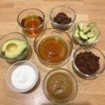 Food-based face masks made from honey, cacao, and avocado displayed in bowls on a wooden table, suitable for skin care.