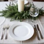 Minimalist holiday meal table setting with simple white plate, silverware, greenery, and candles