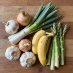 Assorted prebiotic foods such as garlic, onions, leeks, asparagus, and bananas arranged on a rustic wooden surface, ready for cooking.