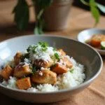 A close-up of a Coconut Chicken Rice Bowl presented on a cozy home dining table, featuring cooked chicken, white rice, shredded coconut, and fresh herbs, with soft natural lighting and a warm, inviting home setting.