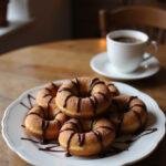 A cozy home setting showing golden churro cruffins on a white plate, coated with cinnamon sugar and drizzled with chocolate sauce, served with a cup of coffee on a wooden table.