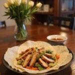 Realistic image of Easy Moo Shu Chicken served on a plate in a cozy home setting, showing sliced chicken with shredded vegetables and pancakes, with warm lighting and home decor background.