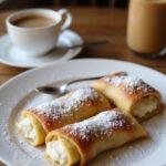 Italian Cream Stuffed Cannoncini on a white plate in a cozy kitchen scene, showcasing golden cannelloni filled with creamy filling, decorated with powdered sugar, with warm lighting and a coffee cup nearby.