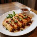 Close-up of crispy oven-fried feta cheese rolls drizzled with chili honey, served on a white plate in a cozy home dining setting, garnished with herbs and accompanied by a small bowl of chili honey.