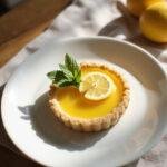 A lemon tart with glossy lemon glaze and flaky crust, garnished with lemon twist and fresh mint, displayed on a white plate in a cozy home setting with warm lighting.