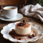 A realistic scene of coffee mousse served on a plate with cocoa powder and coffee bean garnish, set in a cozy home environment with a wooden table, blanket, and a cup of coffee nearby, under warm natural light.