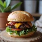 Close-up of a crack burger with a beef patty, melted cheese, crispy bacon, lettuce, tomato, and bun, presented on a wooden surface in a warm home kitchen setting.