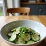 A realistic image of an Asian cucumber salad in a white bowl on a cozy wooden table, featuring fresh sliced cucumbers, sesame seeds, and herbs, in a warm home setting.