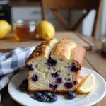 A sliced Lemon Blueberry Yogurt Loaf on a white plate, decorated with blueberries and lemon slices, in a cozy home kitchen setting with warm lighting and rustic decor.