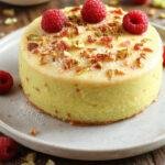 A slice of Pistachio Raspberry Cake showing layers of green pistachio sponge, raspberry filling, and topped with whipped cream and fresh raspberries.