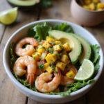 A realistic image of a shrimp and avocado bowl with mango salsa and lime-chili sauce, styled on a cozy wooden table with natural lighting and fresh ingredients around it.