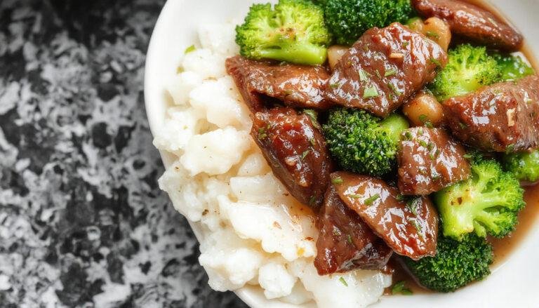 A close-up of a steaming bowl of beef and broccoli stir-fry featuring tender sliced beef, vibrant green broccoli florets, and a glossy savory sauce. The dish is presented on a rustic wooden table with chopsticks resting on the side, highlighting its hearty and colorful appearance.