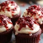A close-up of vampire-themed cupcakes with bright red icing resembling blood, topped with tiny edible fangs and a hint of fake blood sauce, presented on a spooky black platter with Halloween decorations in the background.