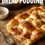 A warm, golden-brown bread pudding served in a rustic white bowl, topped with a dusting of powdered sugar and a drizzle of caramel sauce. The pudding's crispy edges contrast with its soft, custardy interior, garnished with fresh berries and mint leaves. The background features a cozy wooden table with a fork resting on a napkin, evoking a comforting homemade vibe.