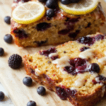 A golden-brown, moist lemon blueberry loaf cake sliced on a white ceramic plate, revealing juicy blueberries embedded throughout, topped with a glossy lemon glaze drizzle; scattered fresh blueberries and lemon zest beside the slice, soft natural light with shallow depth of field.