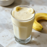A tall glass of creamy yellow Korean banana milk on a light wooden surface, garnished with a banana slice and a splash of vanilla, surrounded by ripe bananas and a drizzle of honey; bright, inviting, and delicious.