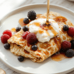 Soft fluffy crepes made from oats and eggs, stacked on a white plate with blueberries and a dollop of yogurt. The batter is pale beige, with a light maple syrup drizzle. Placed on a wooden board with warm morning light and a dusting of cinnamon for a cozy breakfast presentation.