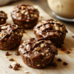 Three glossy no bake peanut butter oat cups sit on a light wooden board, showing creamy peanut butter centers with visible oats and a hint of chocolate drizzle. The cups have a natural, homemade texture and are framed by a clean, bright kitchen setting with soft shadows and warm lighting.